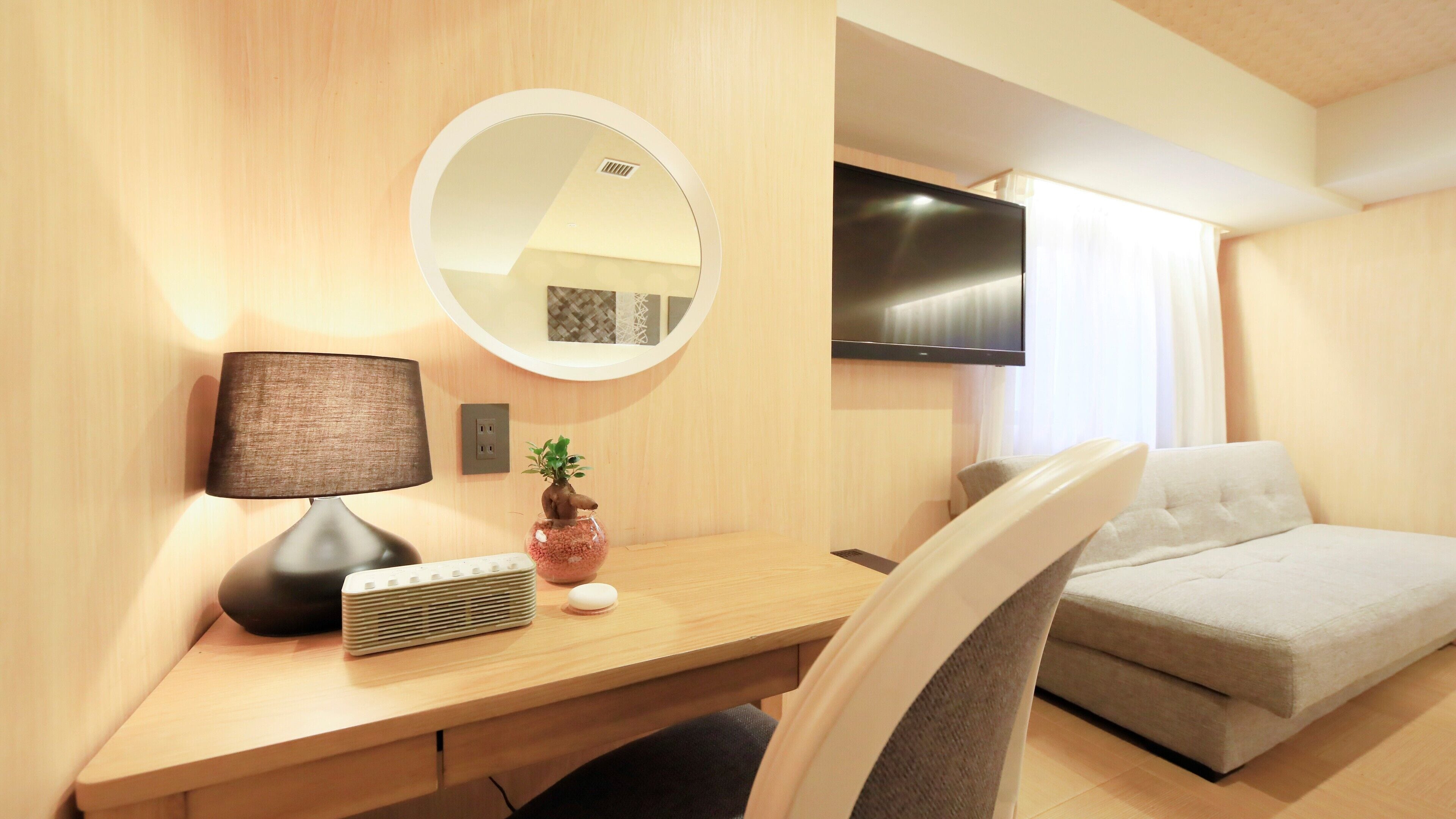 Une chambre d’hôtel avec un bureau en bois, un miroir rond, un téléviseur à écran plat, un canapé et une petite plante en pot.