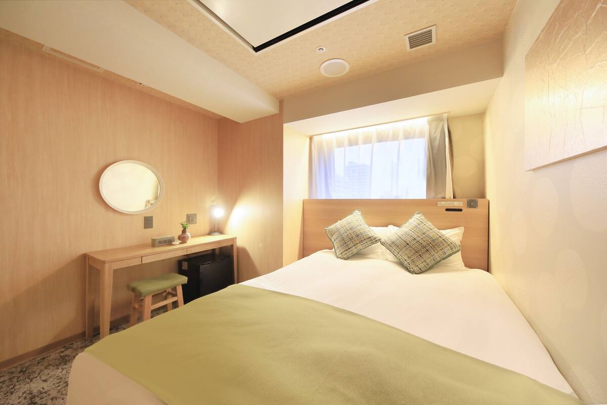 A hotel room with a bed, a desk, a chair, a mirror, and a window with curtains.