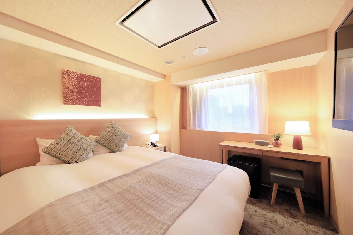 A hotel room with a large bed, a desk, a chair, a lamp, and a window with curtains.