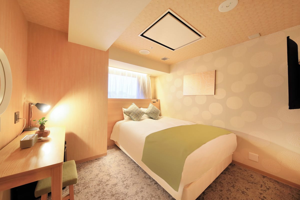 A hotel room with a bed, a desk with a lamp, a small table with a plant, and a window with curtains.