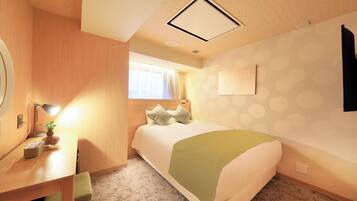 A hotel room with a bed, a desk with a lamp, a small table with a plant, and a window with curtains.