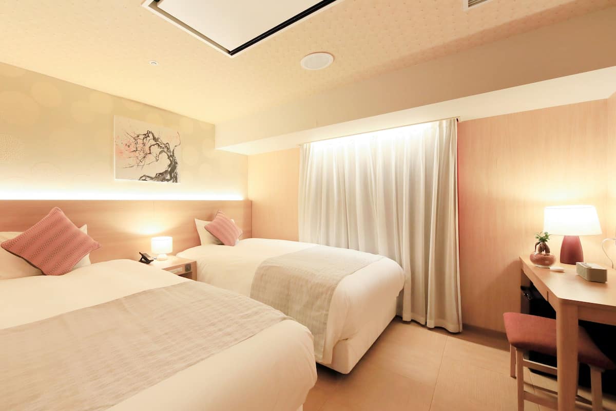 A hotel room with two beds, a desk with a lamp, a chair, and a large window with curtains.