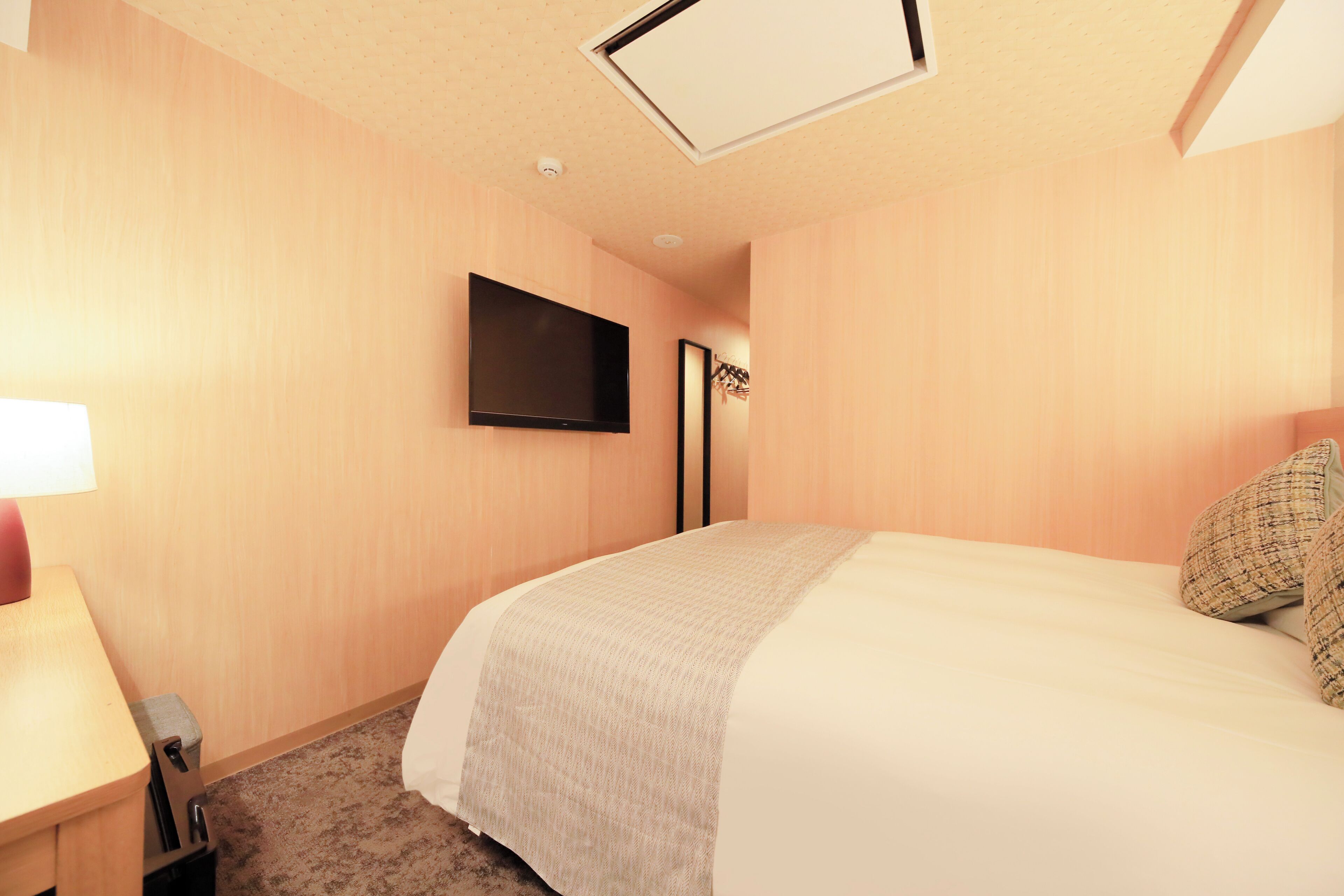 A hotel room with a bed, a flat-screen TV mounted on the wall, a desk with a lamp, and a carpeted floor.