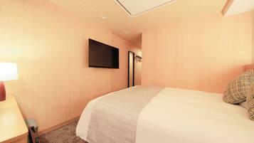 A hotel room with a bed, a flat-screen TV mounted on the wall, a desk with a lamp, and a carpeted floor.
