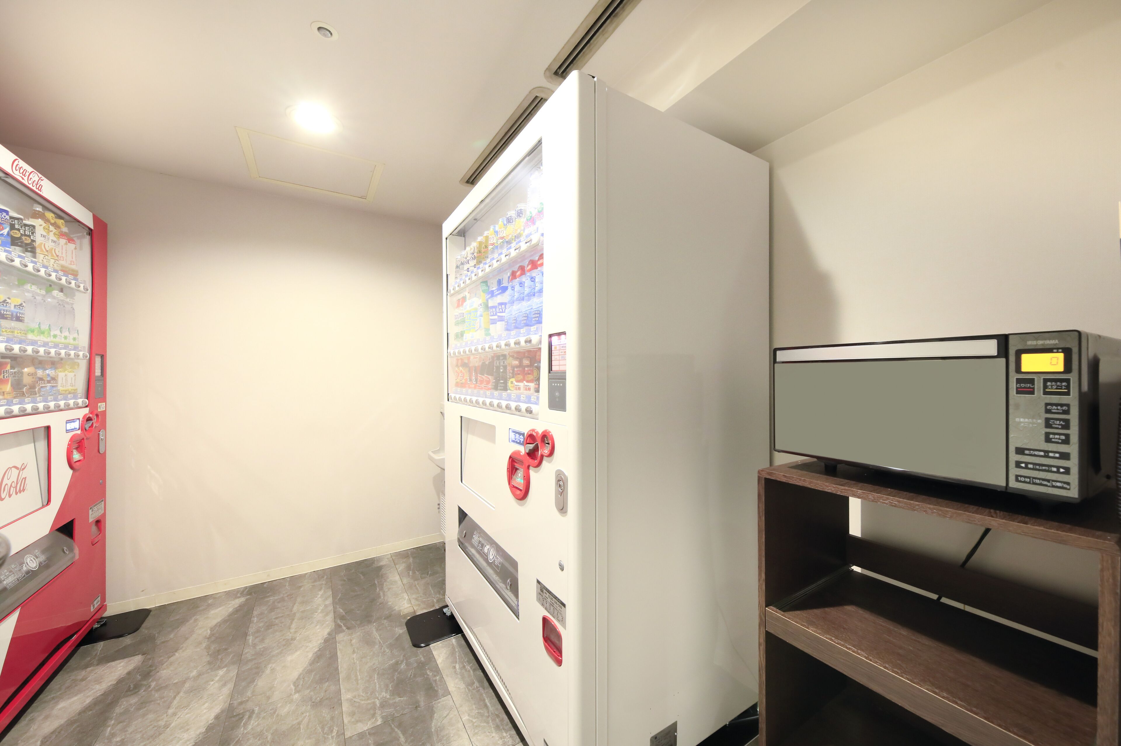 A room with a vending machine stocked with beverages, a microwave, and a wooden shelf.