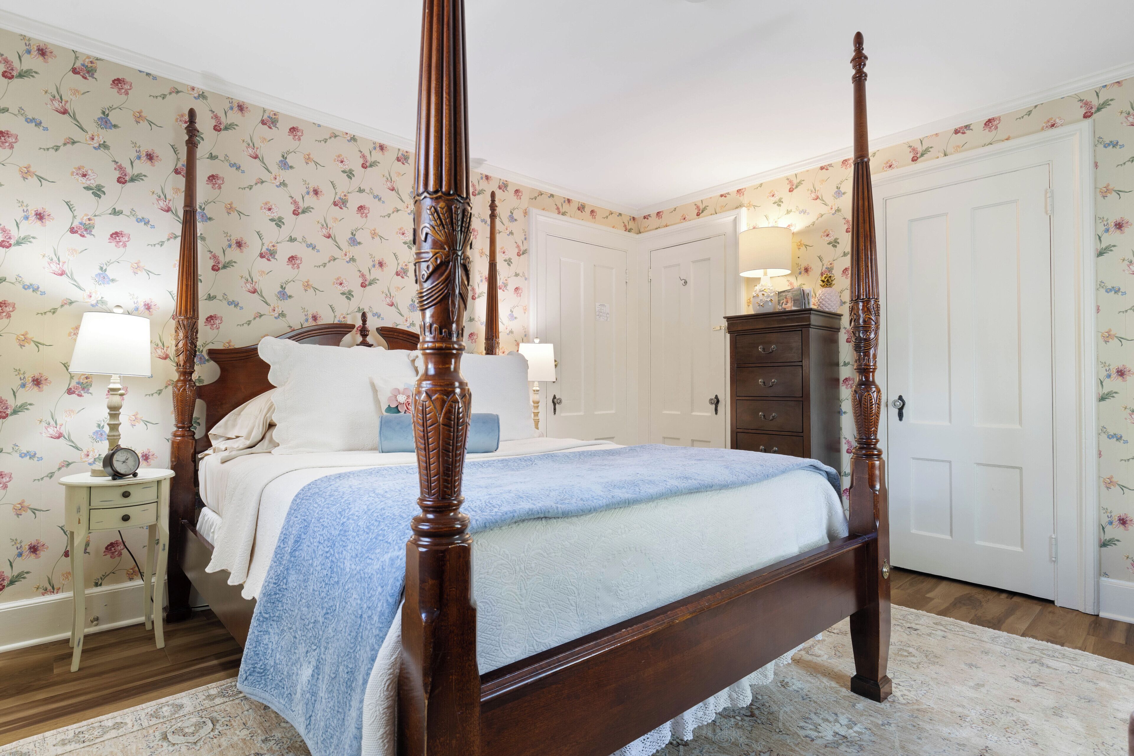 A four-poster bed with a wooden frame and a blue bedspread, a nightstand with a lamp, a dresser, and a door with floral wallpaper.