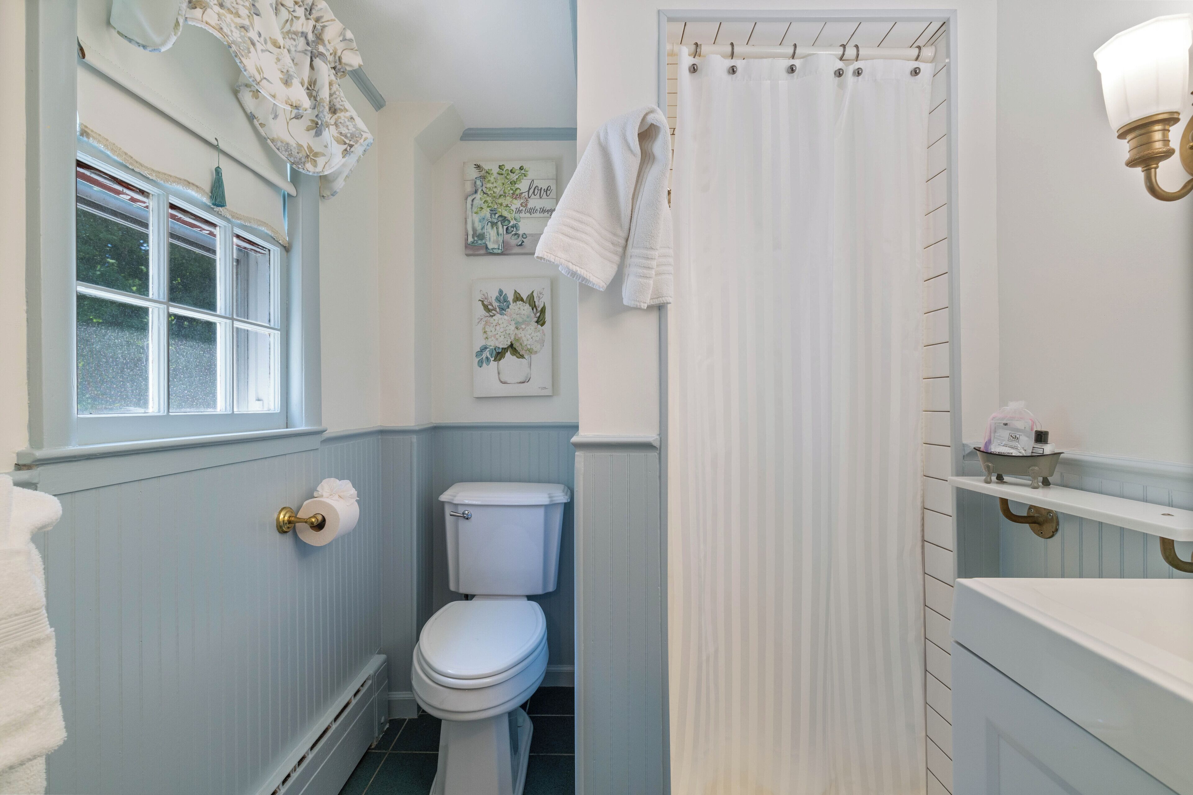 A bathroom with a toilet, a shower curtain, a window with a curtain, and a wall-mounted shelf with toiletries.