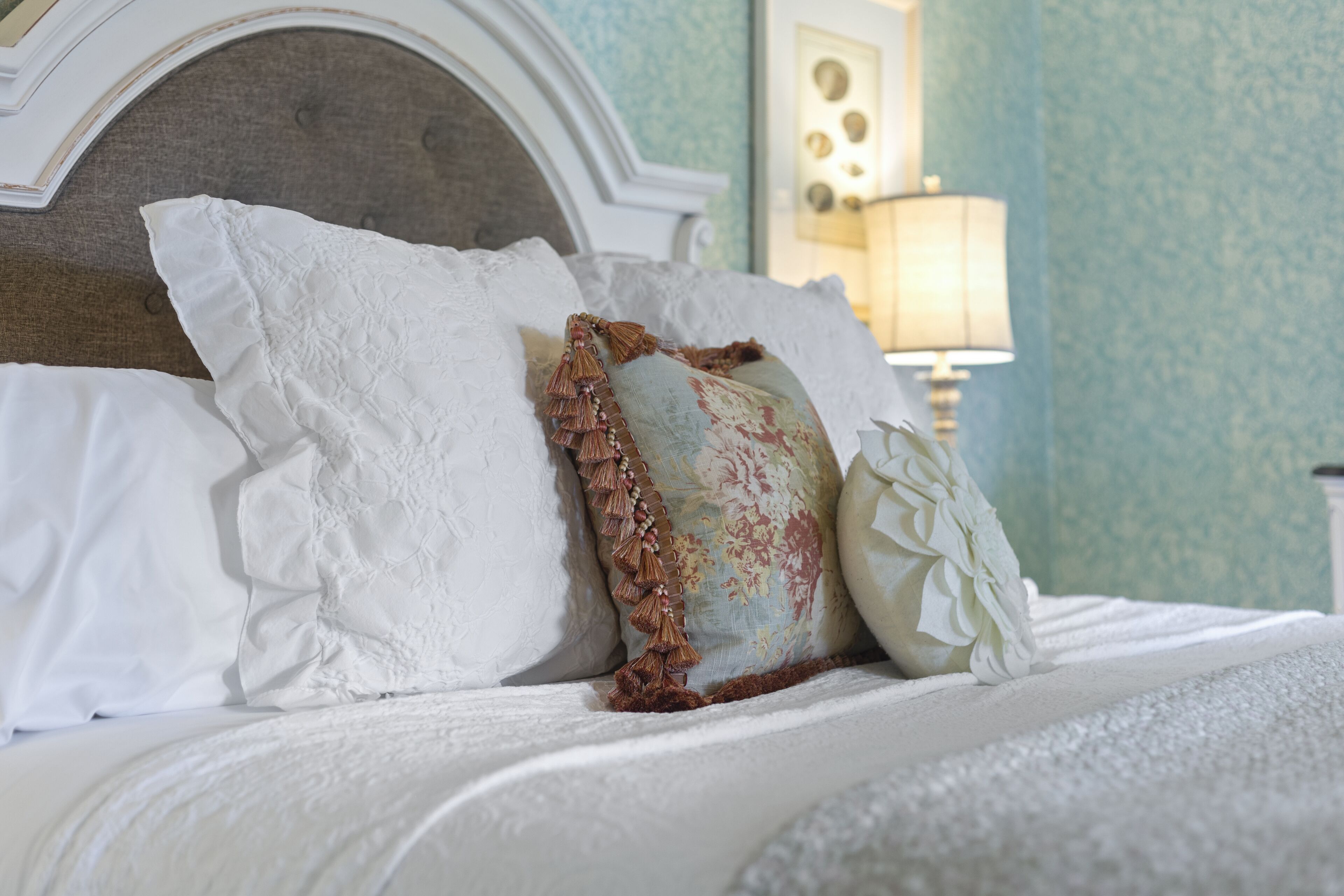 A neatly made bed with white linens and decorative pillows, a classic headboard, and a lamp in the background.