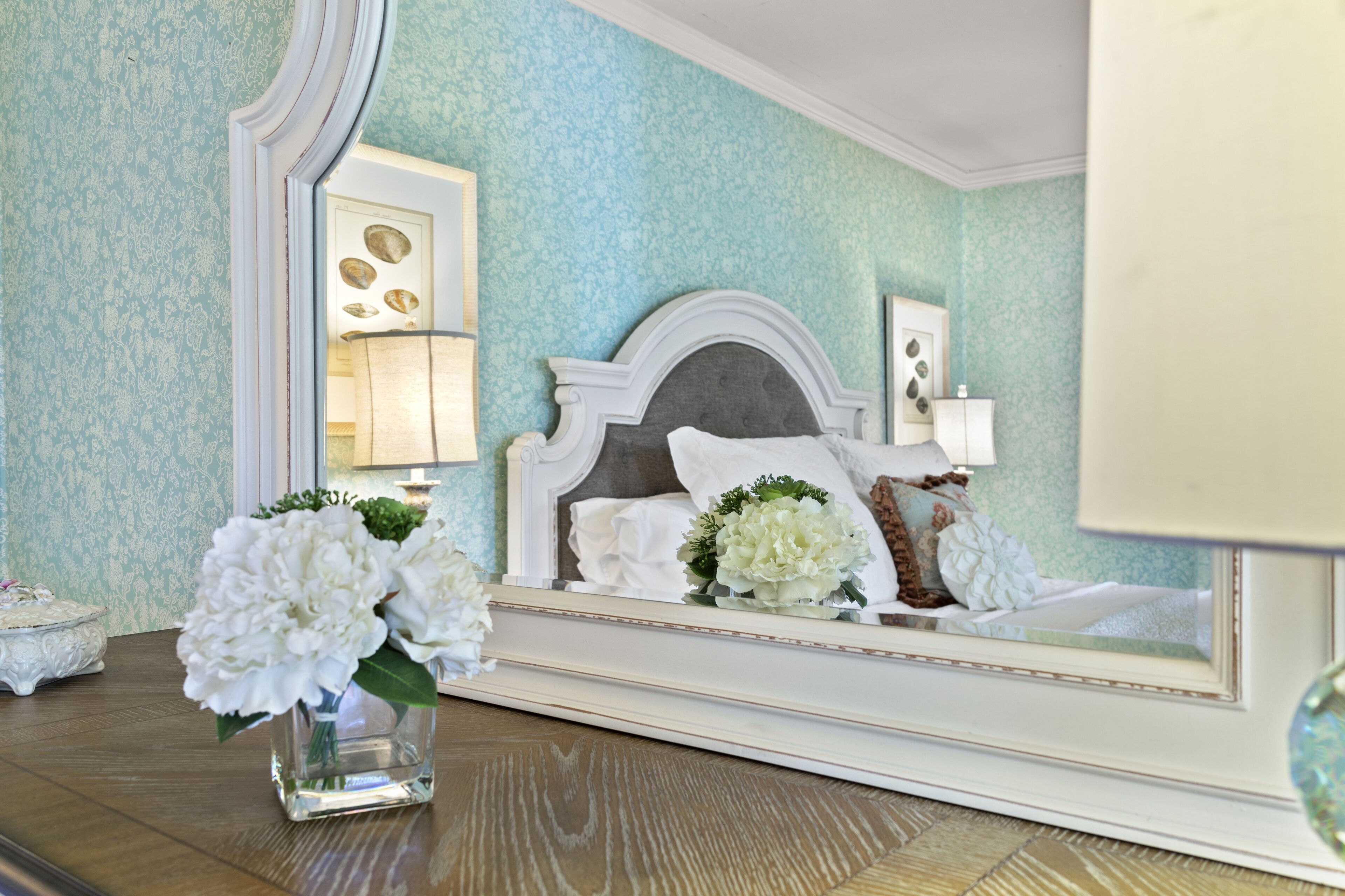 Een slaapkamer met een grote spiegel, een bed met witte lakens, en een vaas met witte bloemen op het nachtkastje.