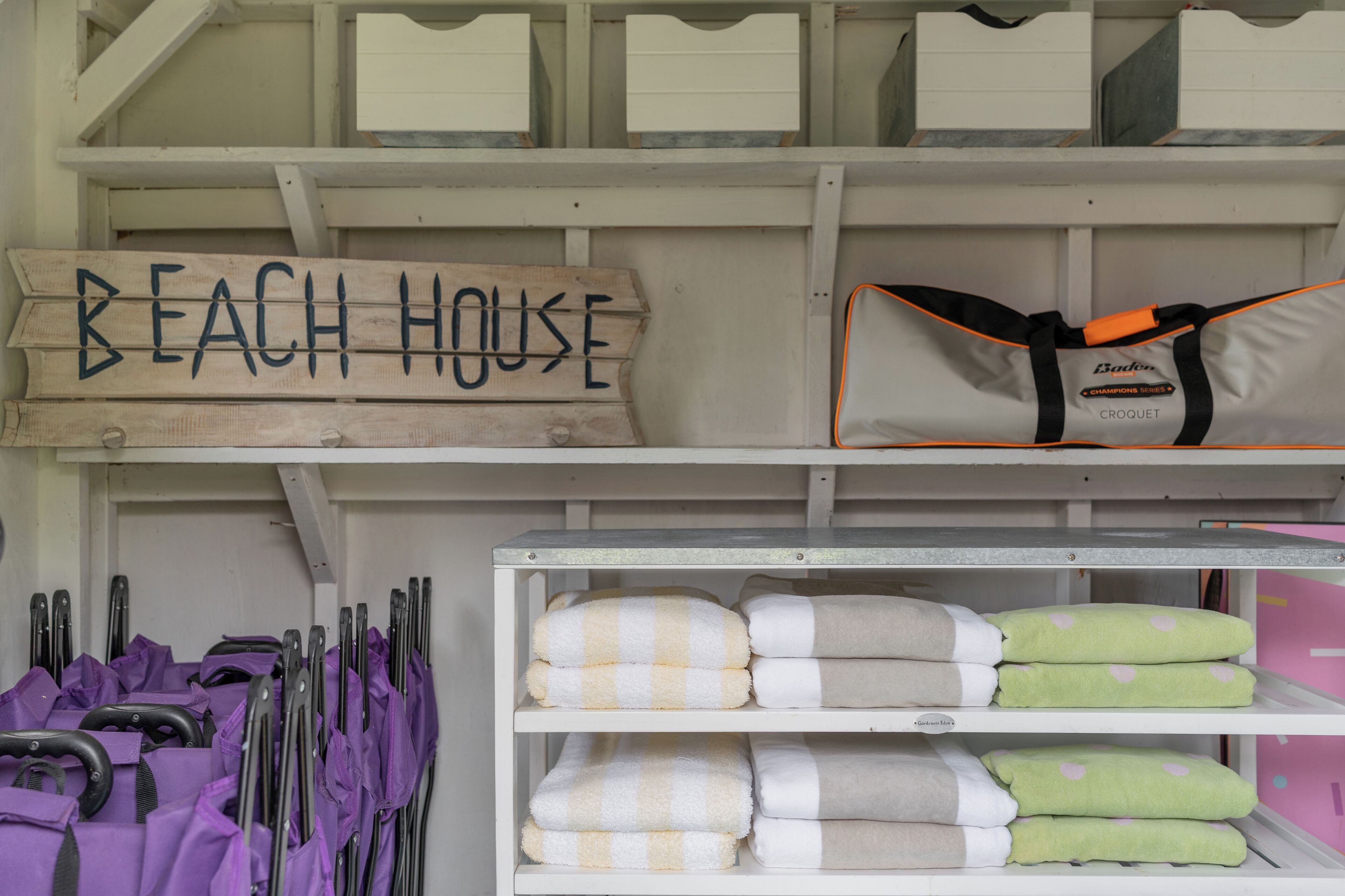 Un área de almacenamiento con un cartel que dice 'BEACH HOUSE', una bolsa de la marca 'Bader' y toallas dobladas con cuidado.
