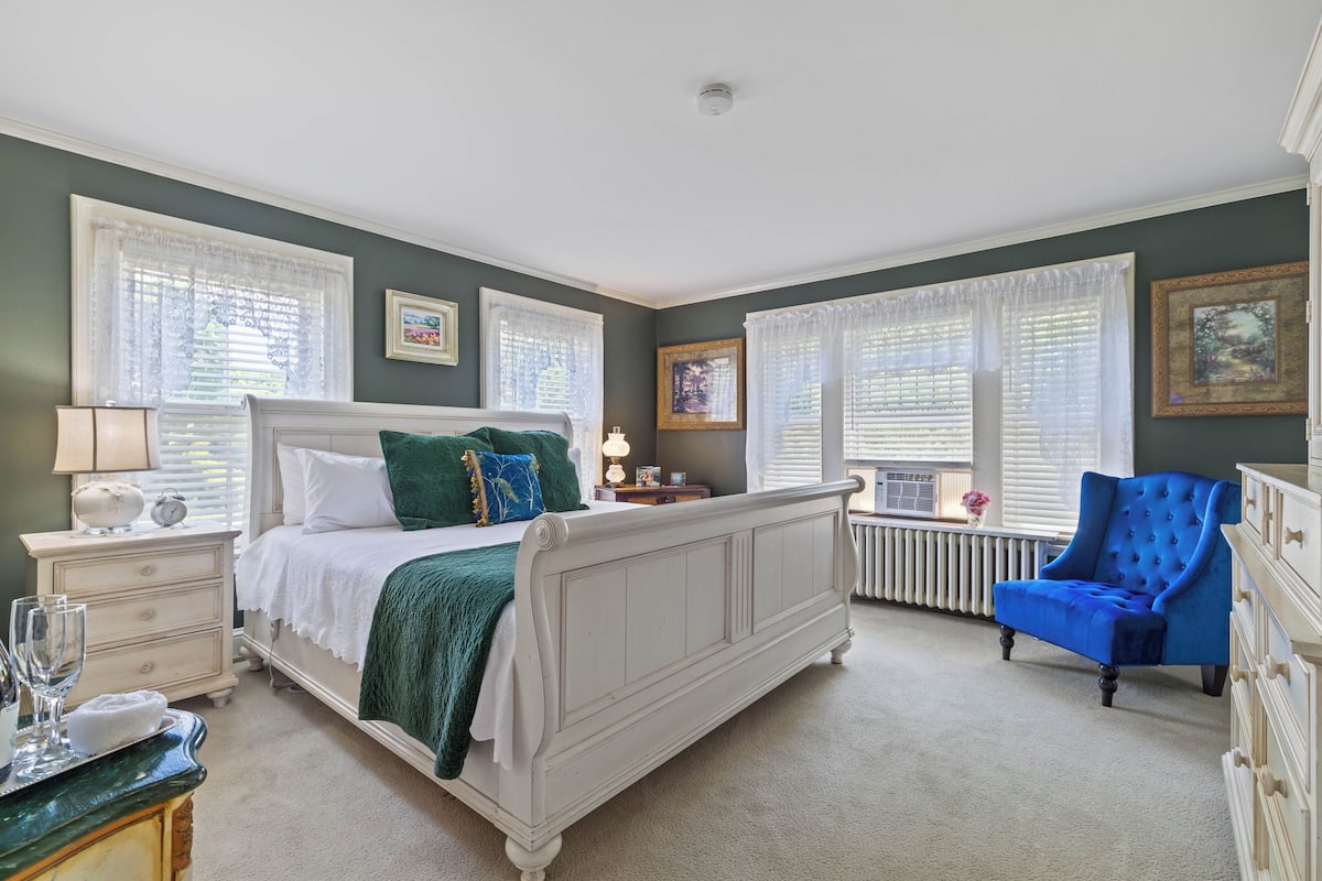 Quarto com cama branca, almofadas verdes, poltrona azul e cômoda.