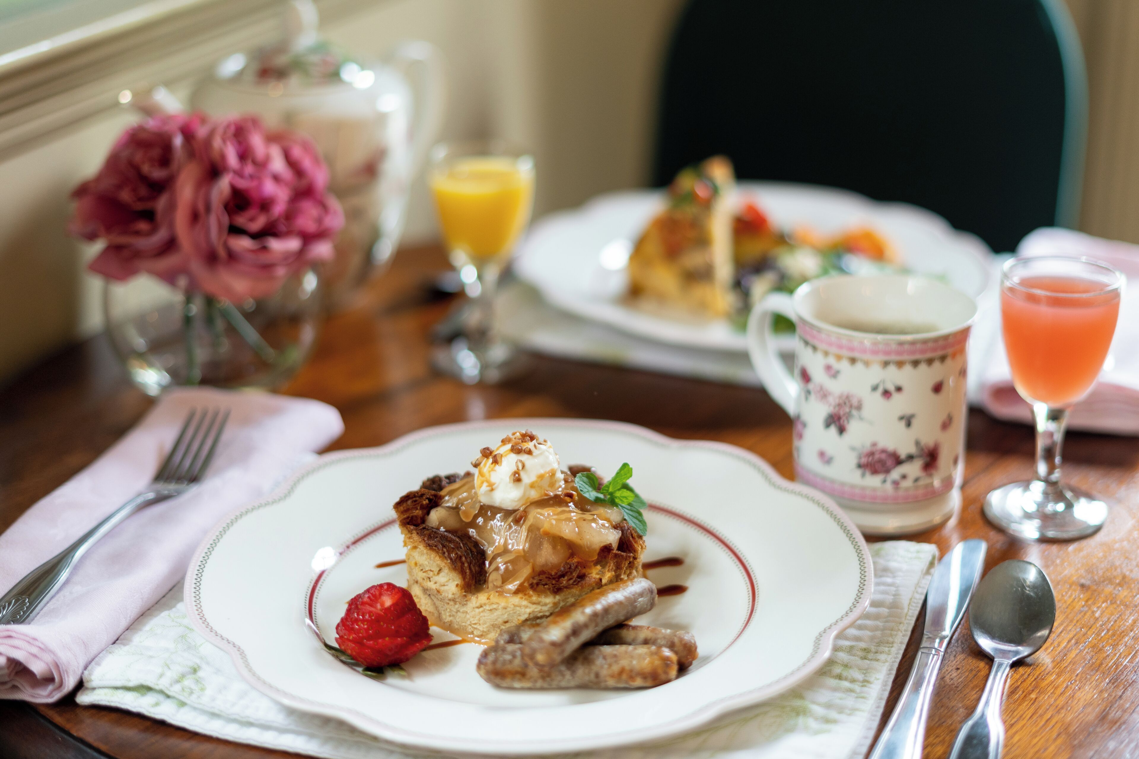 A breakfast setting with a plate of pancakes, sausage, and a strawberry, accompanied by a glass of orange juice and a cup of tea.