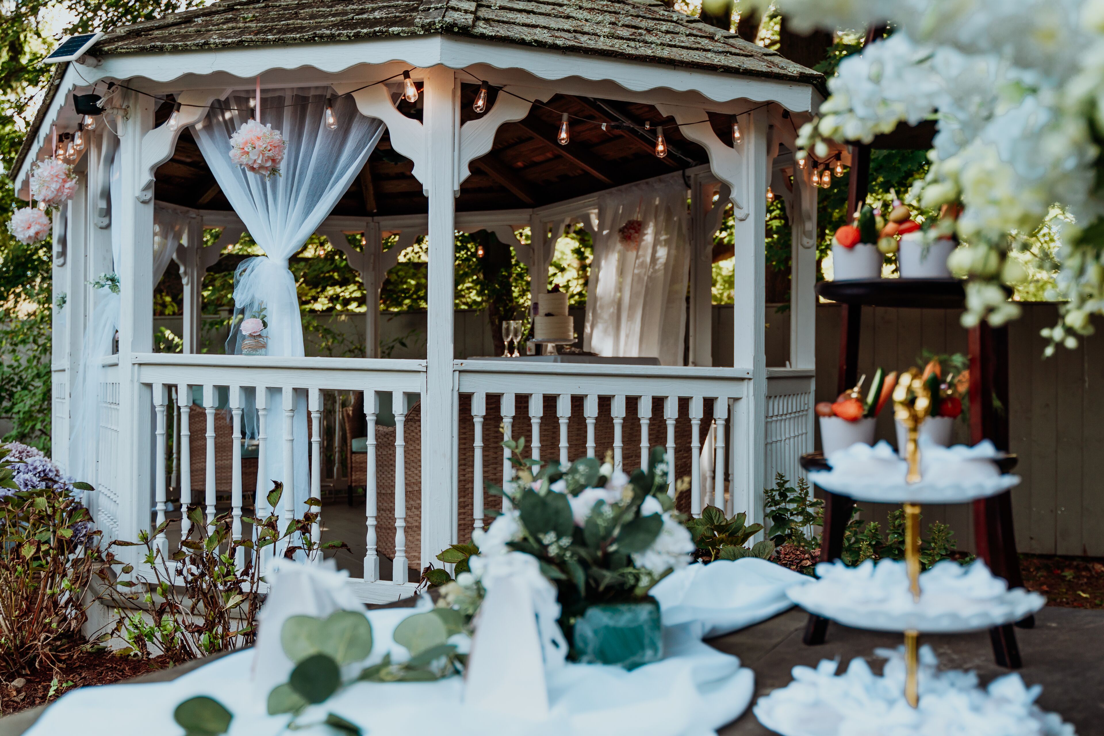 A gazebo with a white exterior and a wooden roof, surrounded by greenery and white floral decorations.