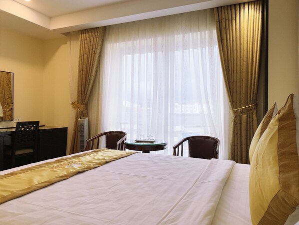 A hotel room with a large bed, two chairs, a small table, and a window with sheer curtains.