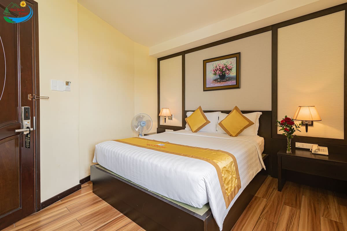 A hotel room with a bed, bedside tables, a nightstand, a wall-mounted fan, a painting, and a wooden floor.