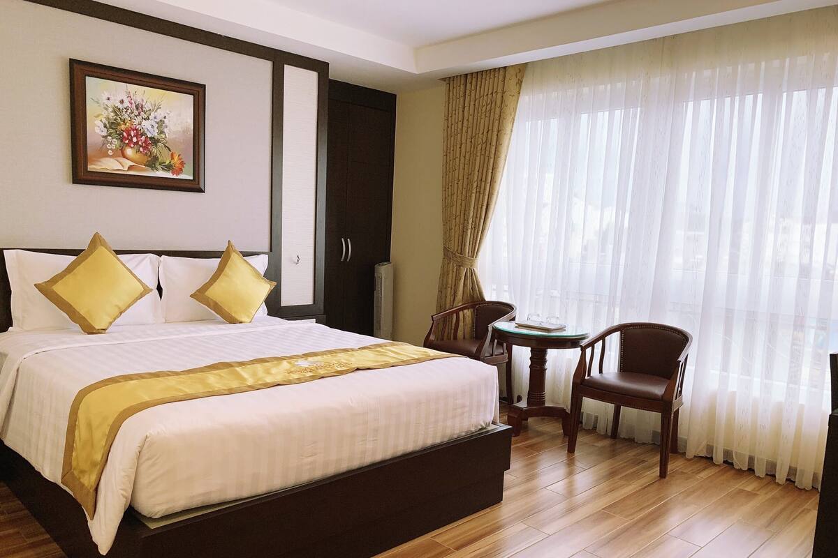A hotel room with a large bed, two chairs, a small table, and a window with sheer curtains.