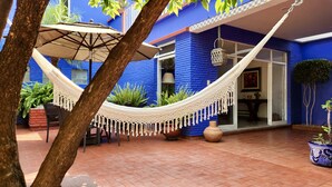 A hammock set up in a courtyard with a blue building, a potted plant, and a tiled floor. - Casa Tamayo (Mexico City)