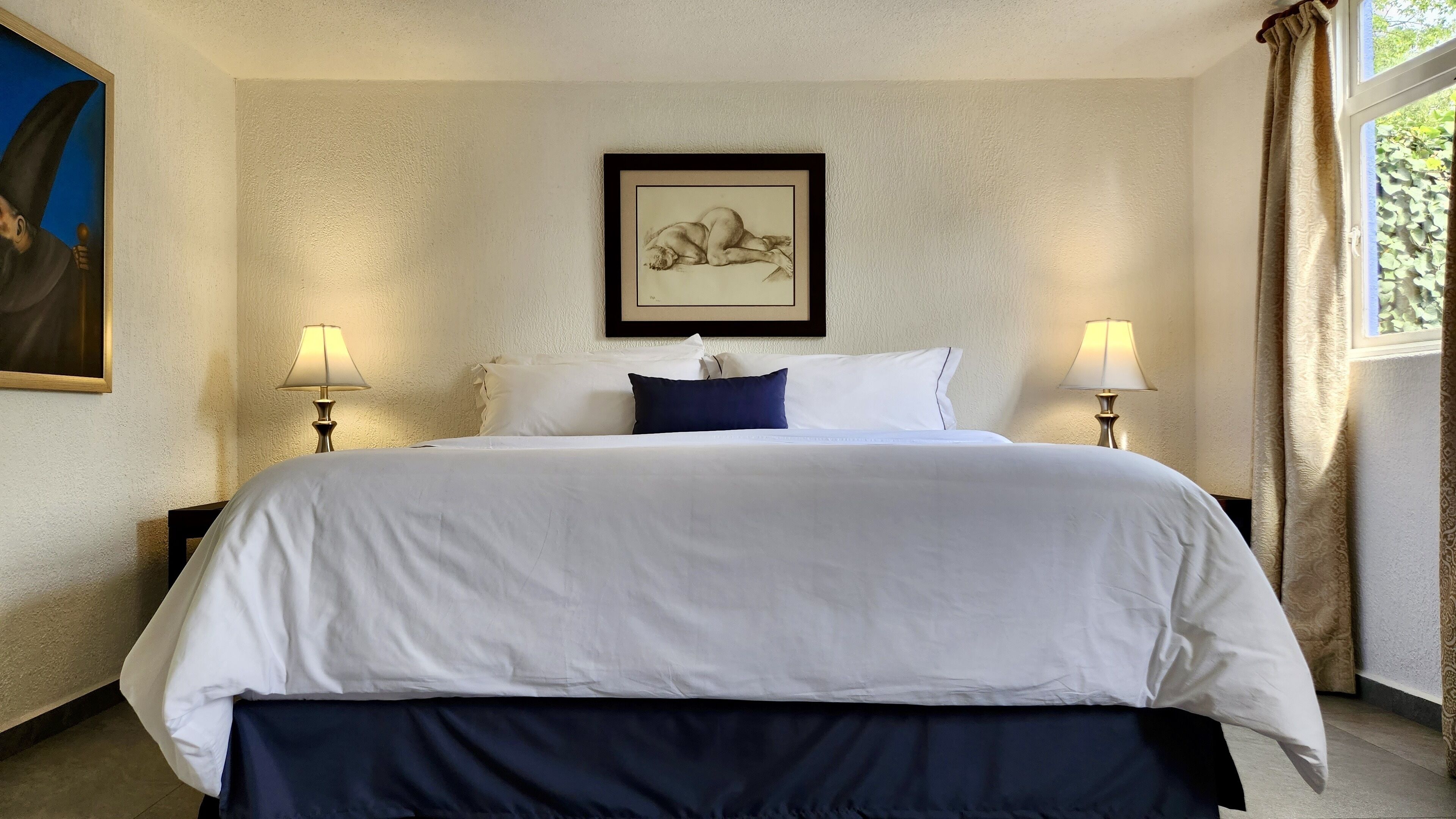A hotel room with a large bed, two bedside lamps, a framed artwork on the wall, and a window with curtains.
