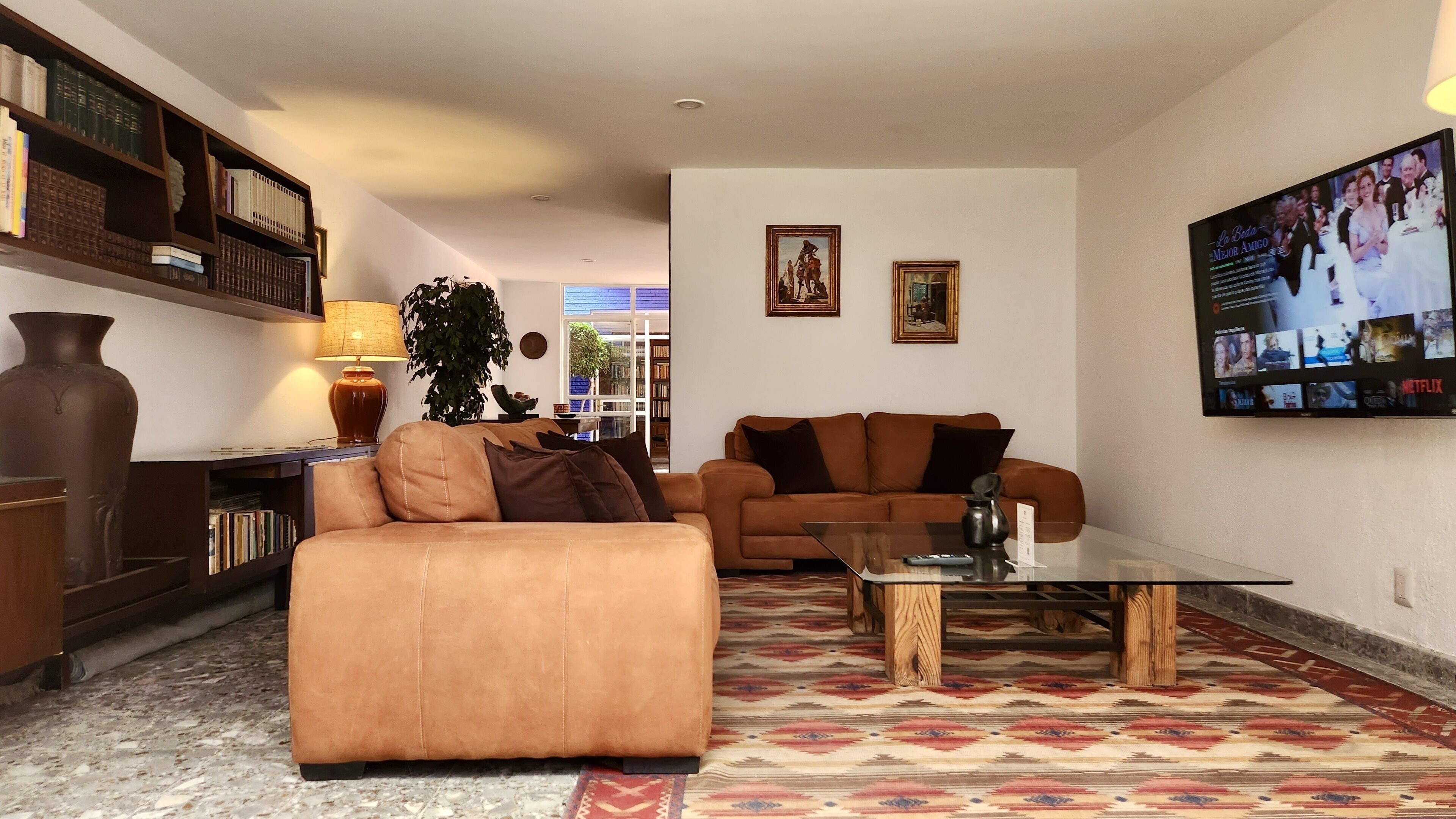 A living room with a brown sofa, a glass coffee table, a bookshelf, and a TV mounted on the wall displaying a movie.
