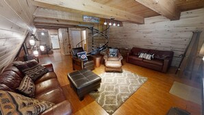 Ein gemütliches Blockhaus-Interieur mit einem Wohnzimmer, das Ledersofas, einen gemusterten Teppich und eine Treppe in einen Küchenbereich zeigt.