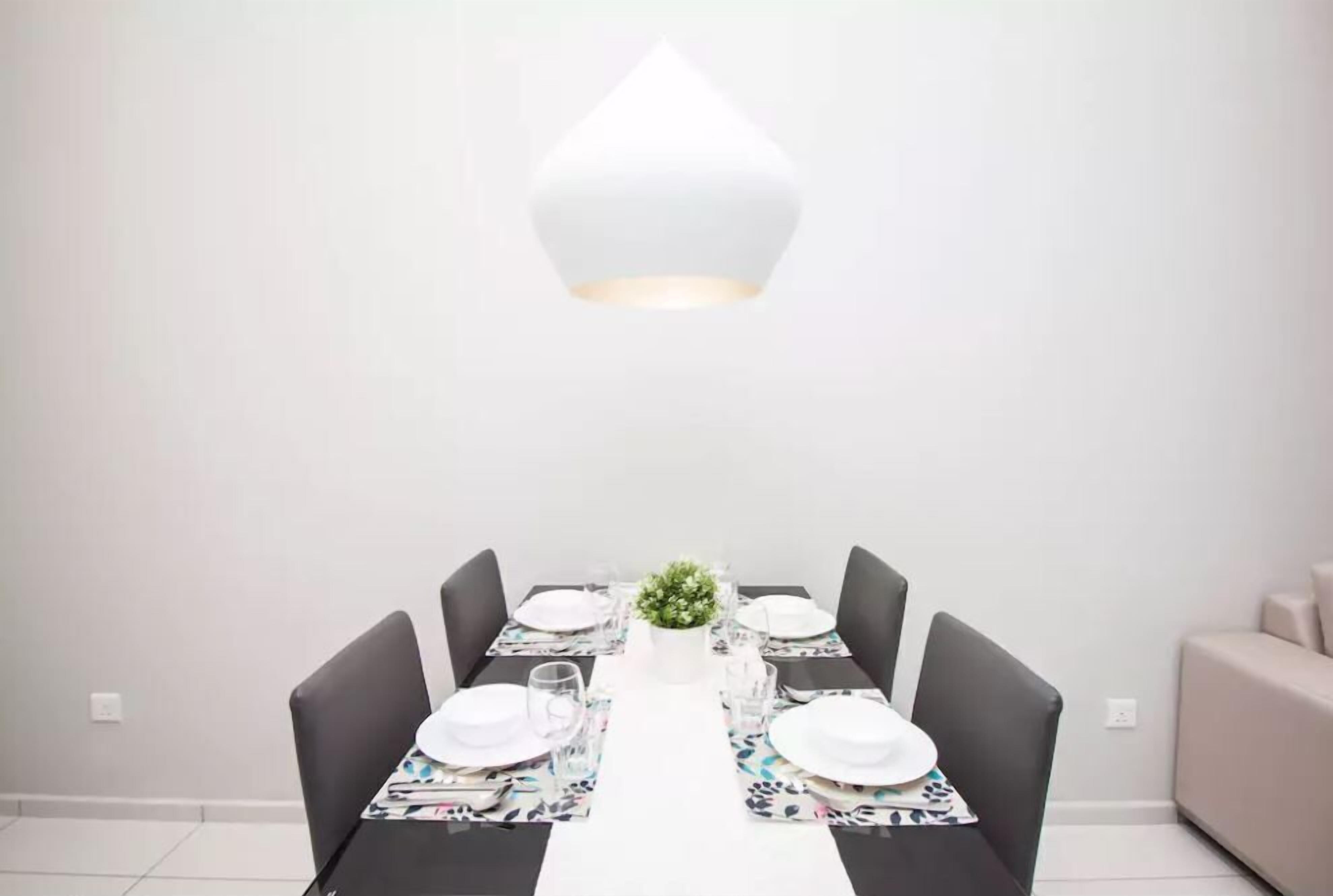 Un comedor con una mesa blanca para cuatro personas, rodeada de sillas oscuras, una planta central y una lámpara colgante moderna.