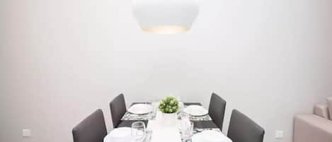 Un comedor con una mesa blanca para cuatro personas, rodeada de sillas oscuras, una planta central y una lámpara colgante moderna.