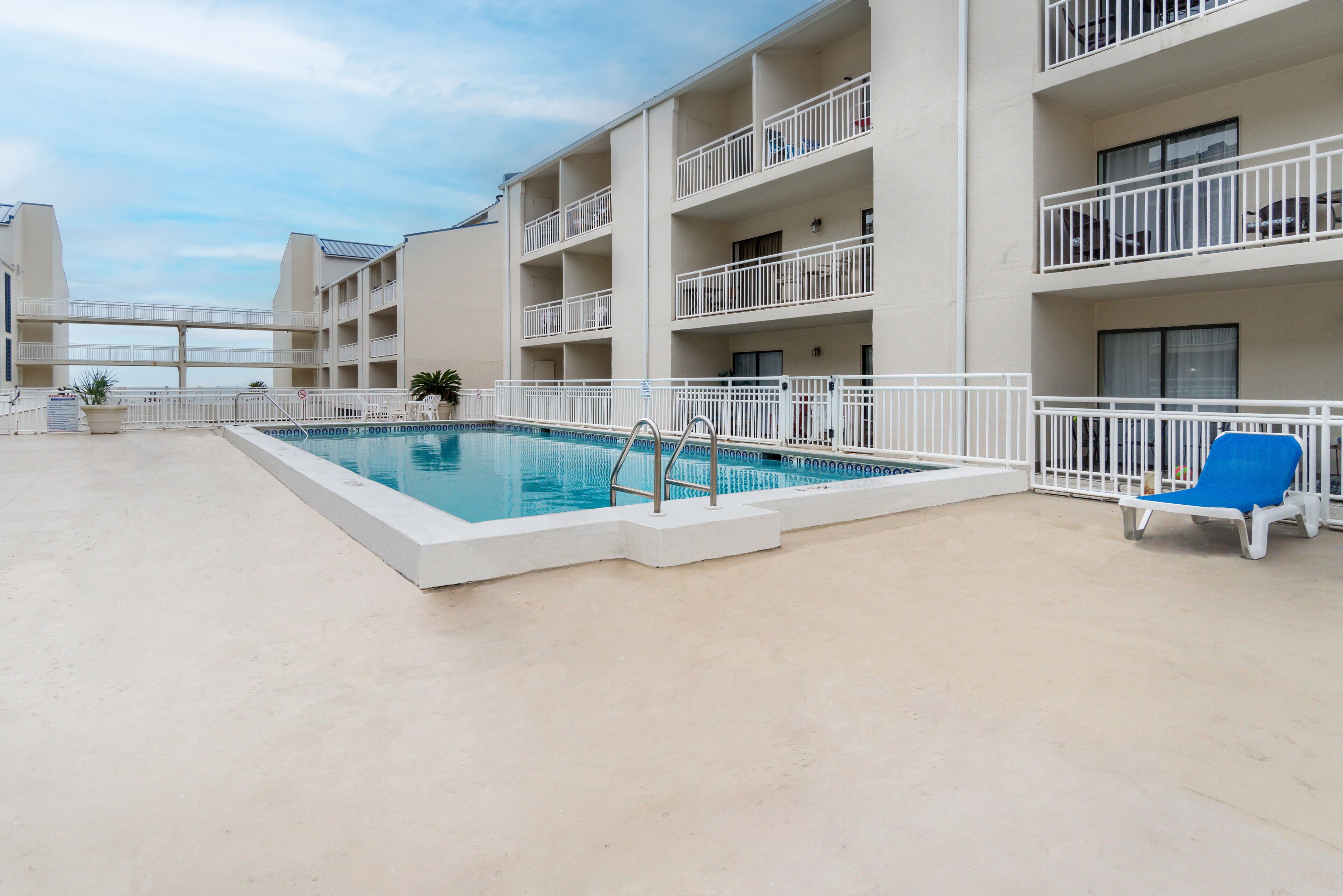 A swimming pool with a white deck, surrounded by a white railing, and a blue lounge chair.