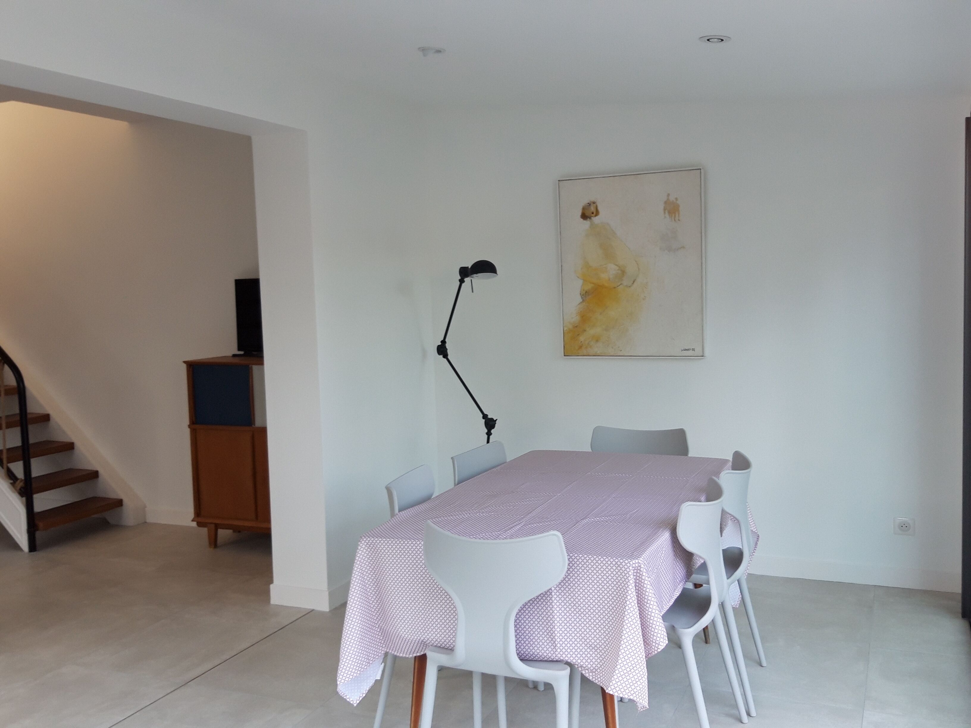 A dining area with a table set for four, white chairs, a black floor lamp, and a painting on the wall.