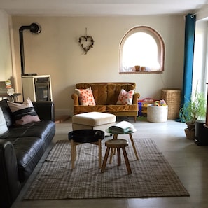 A living room with a brown sofa, a black leather couch, a small wooden stool, and a white ottoman. There is a window with a blue curtain, a heart-shaped decoration on the wall, and a basket of toys on the floor. - Spacious Waterfront House with Jacuzzi for Families and Friends (Nargis)