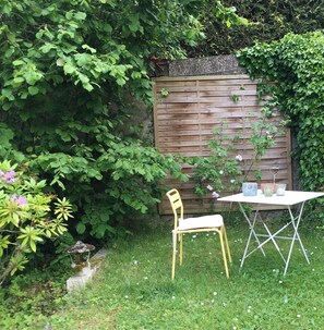 Un jardin avec une clôture en bois, une chaise jaune et une table blanche ornée d’un vase et d’un verre.