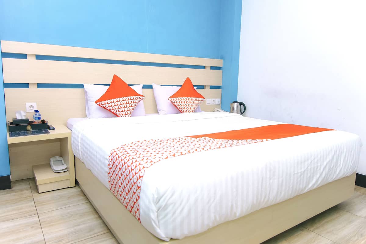 A neatly made bed with white and orange bedding, a wooden headboard, and a nightstand with a phone and a water bottle.