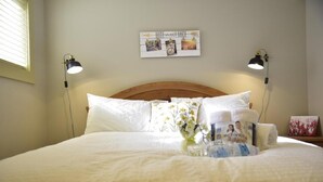 A neatly made bed with white linens, two bedside lamps, a wall decoration, and a small table with a vase of flowers and a water bottle. - Calgary Cozy Home (Calgary)