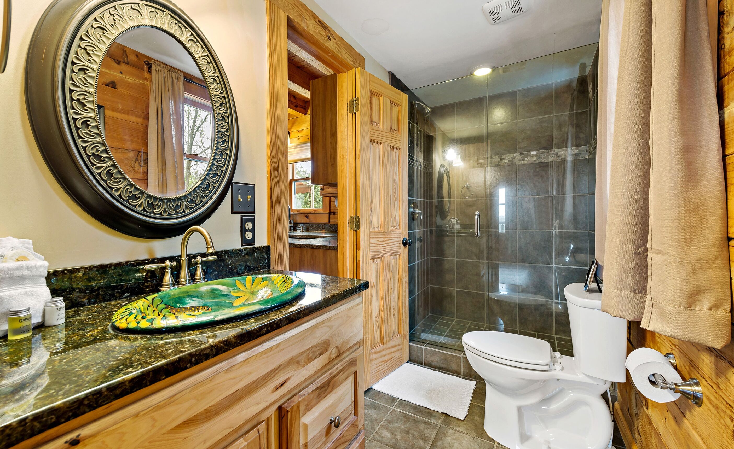 A bathroom with a wooden vanity, a green ceramic sink, a large mirror, and a glass-enclosed shower.