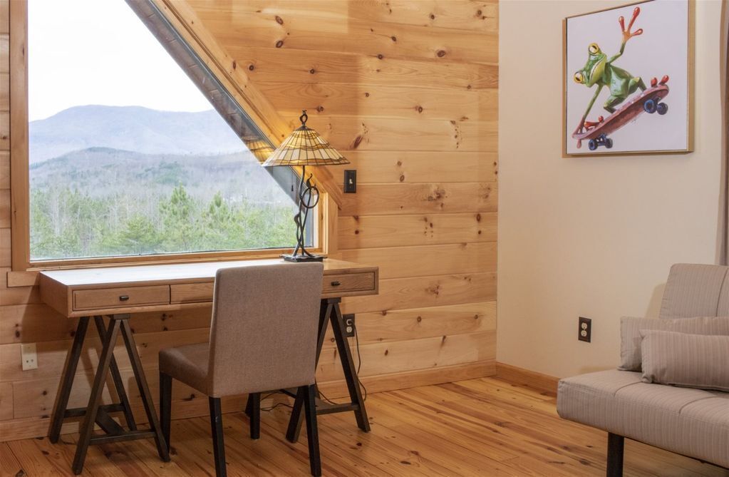 A wooden cabin room with a desk, chair, and a large window overlooking a forested landscape.