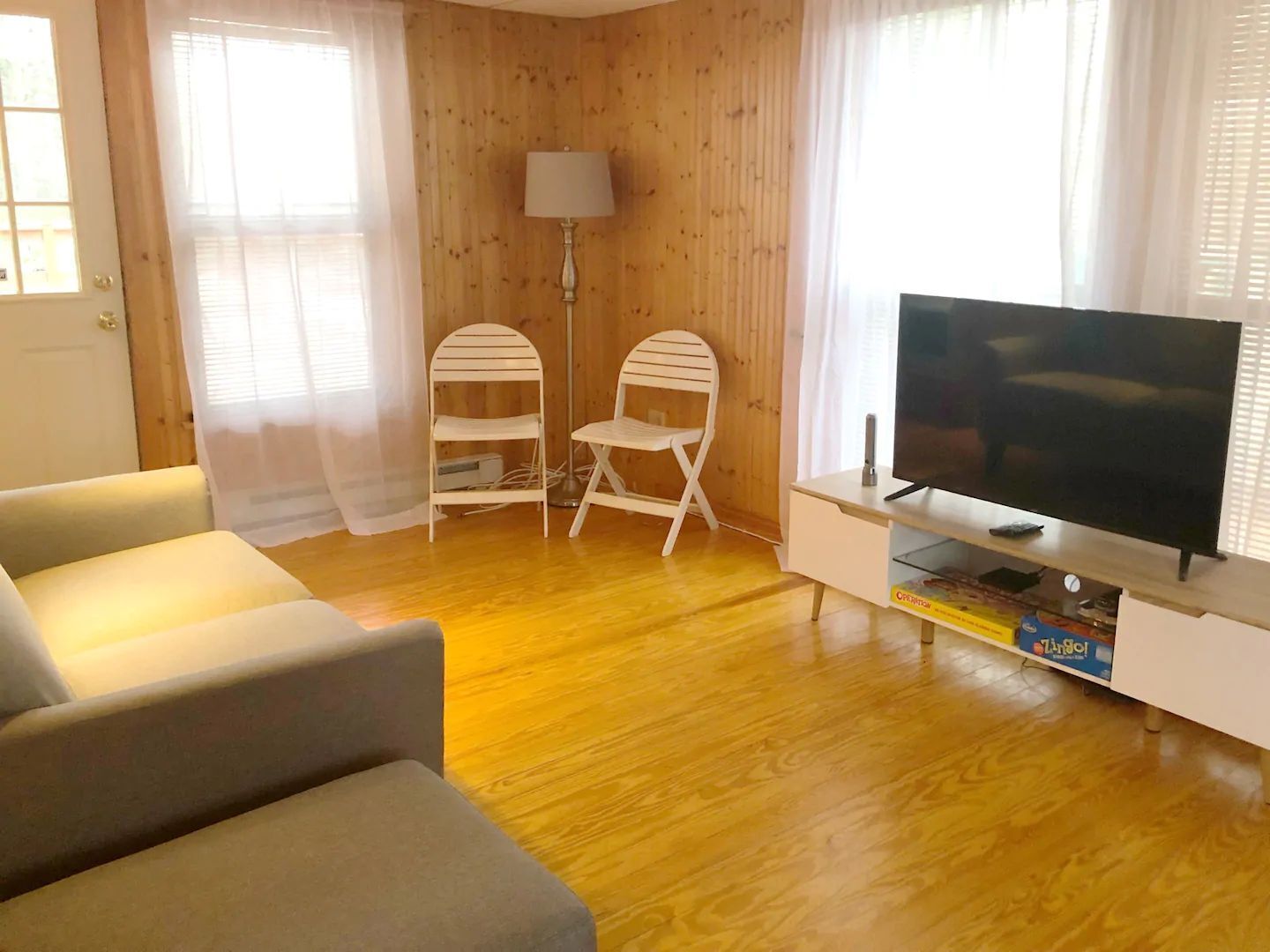 Una sala de estar con un sofá, un televisor sobre un mueble bajo, una lámpara y dos sillas blancas.