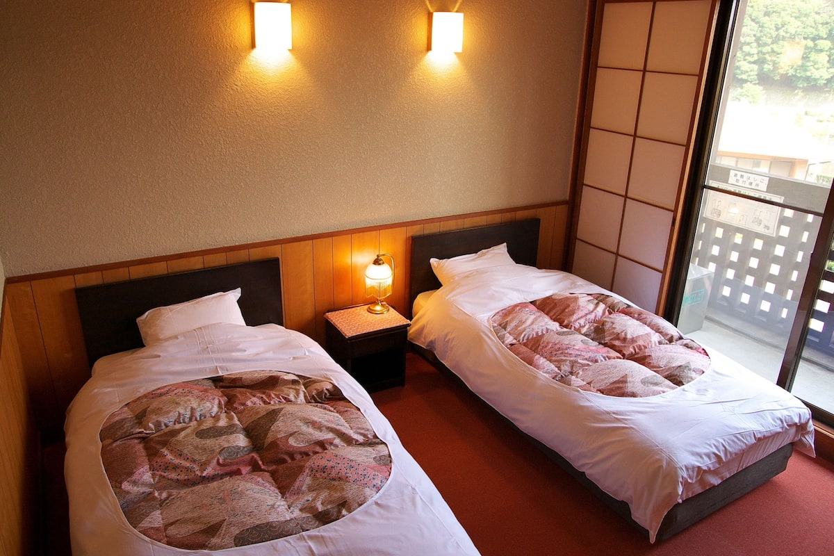 A hotel room with two beds, a nightstand with a lamp, and a view of the outside through a large window.