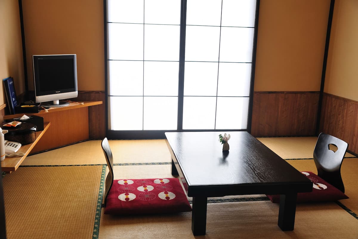 A traditional Japanese room with tatami flooring, a low table, a chair, and a television on a shelf.