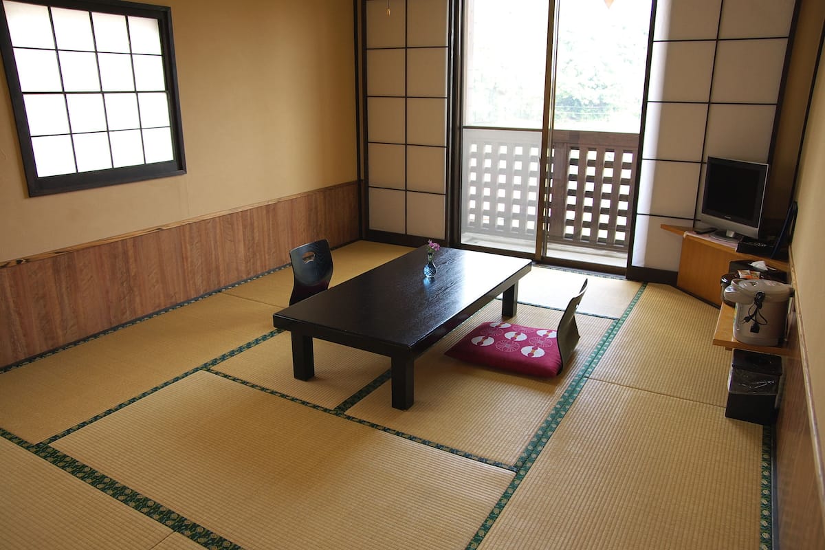 A tatami mat room with a low table, a chair, a small table with a vase, a television, and a sliding door.