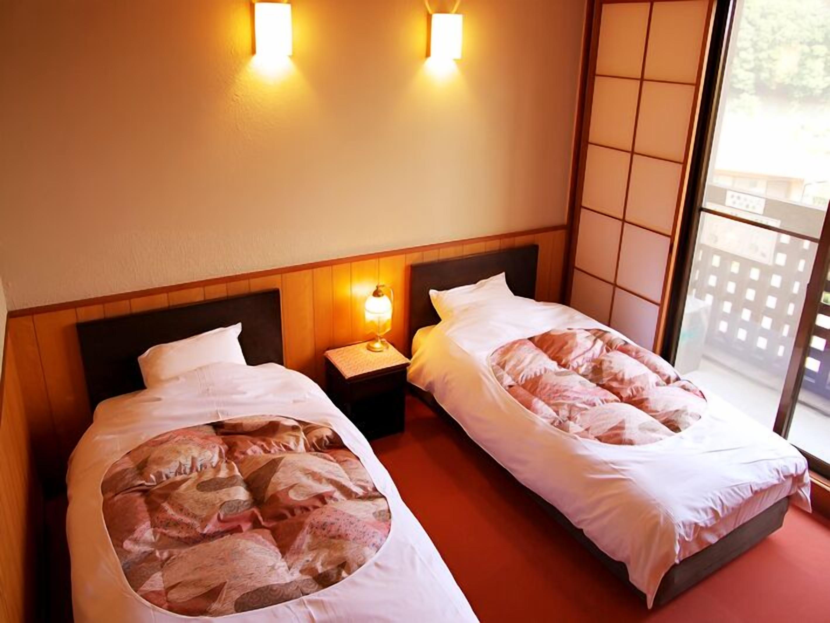 Une chambre d’hôtel avec deux lits, une table de chevet avec une lampe et une porte-fenêtre coulissante donnant sur l’extérieur.
