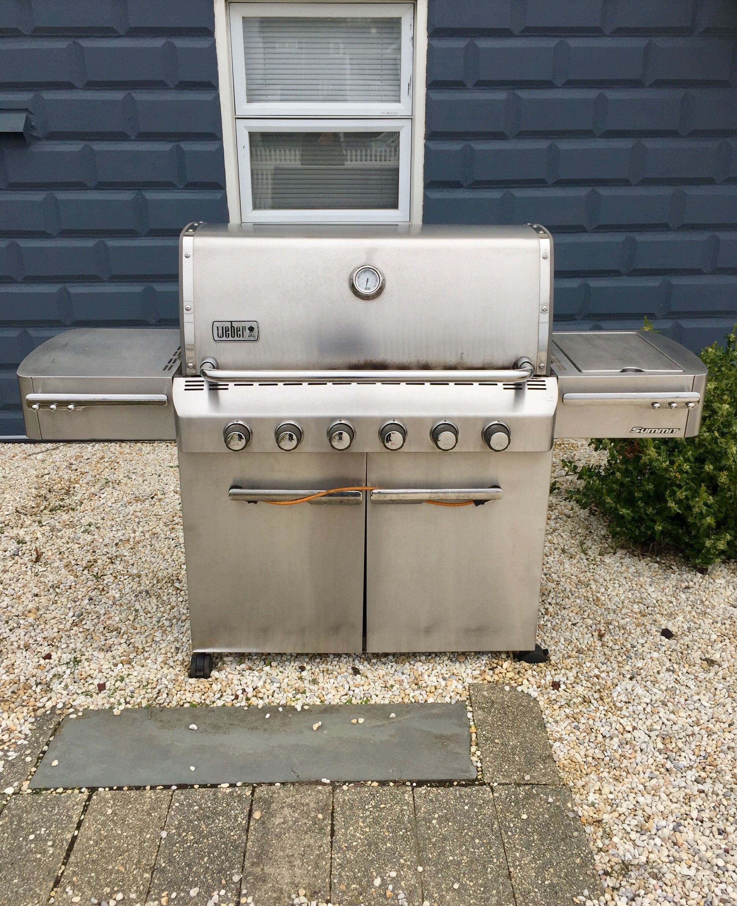 Ein Weber-Grill aus Edelstahl mit integriertem Thermometer und mehreren Brennern, aufbefindlich auf einer Schotterfläche vor einer blauen Wand.