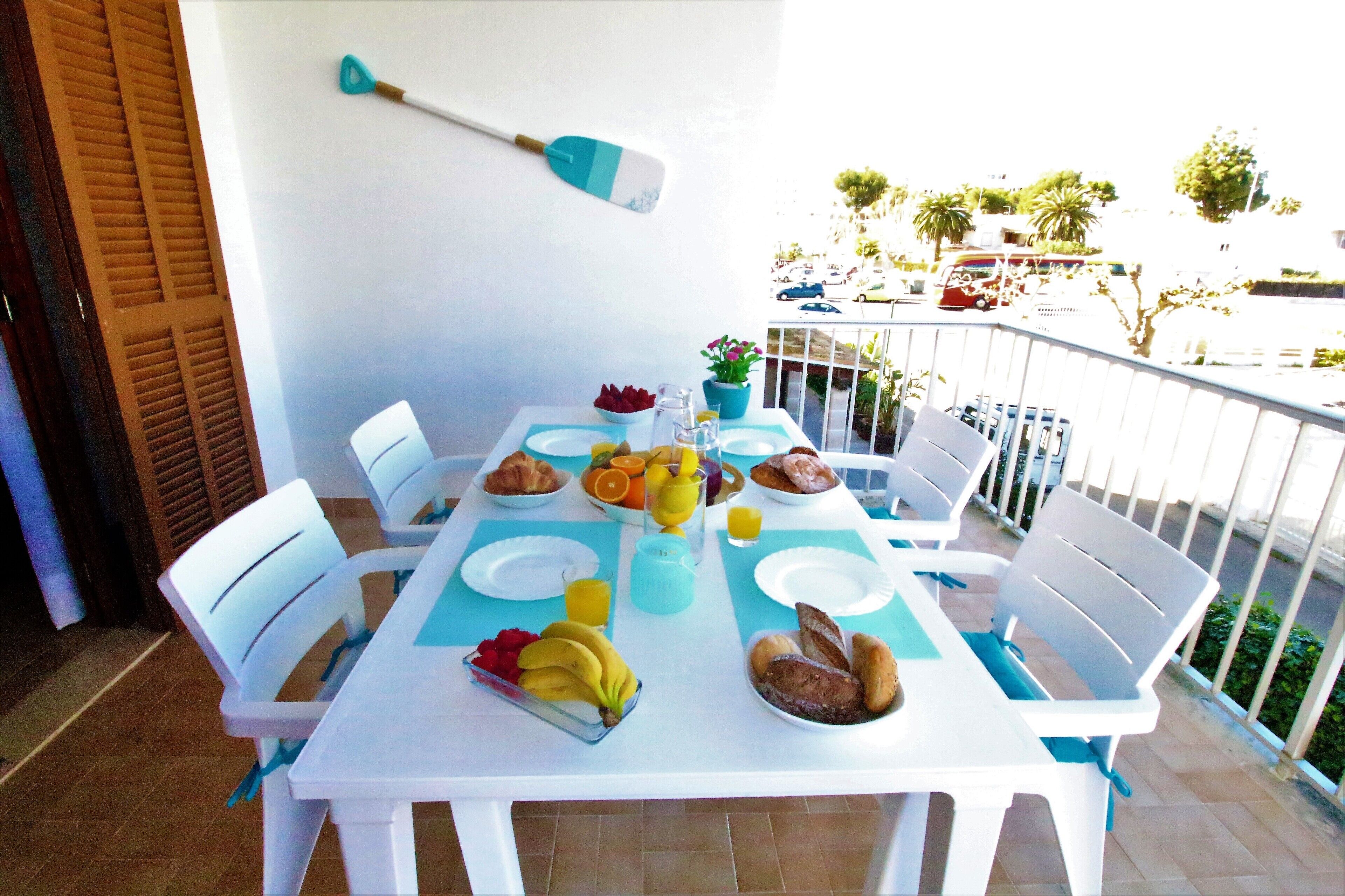 Ein weiß gedeckter Tisch mit Frühstücksleckereien, darunter Früchte, Brot und eine Kanne Orangensaft, auf einem Balkon mit Blick auf parkende Autos und Palmen.