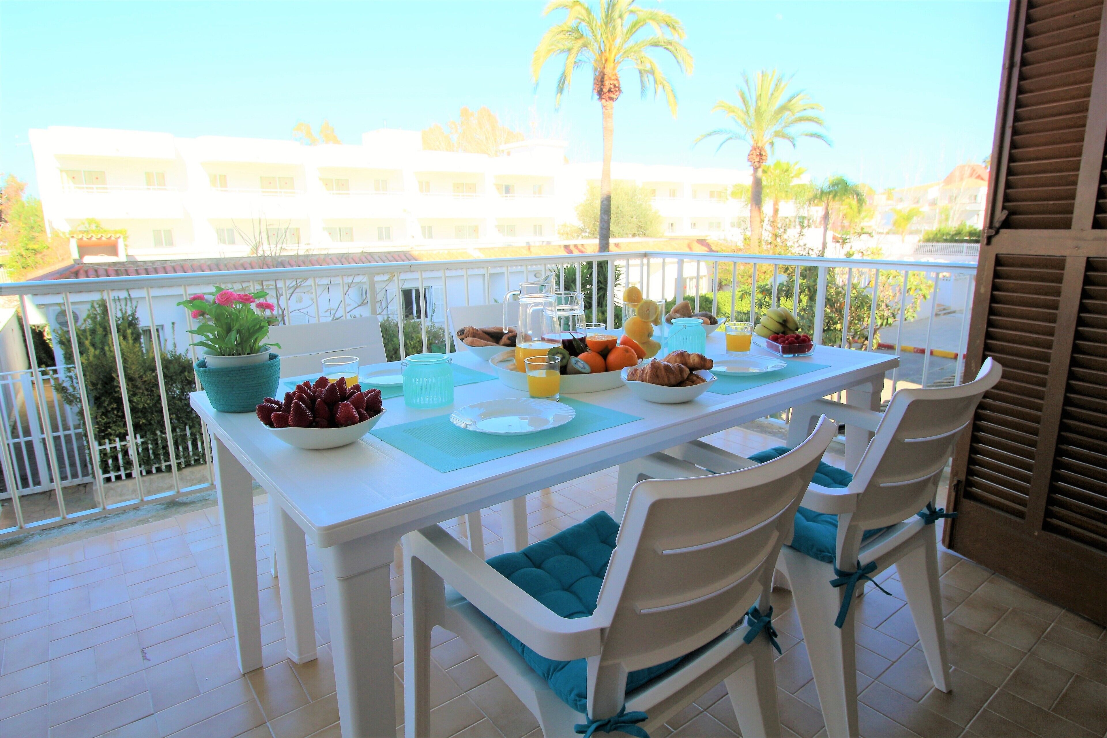 A balcony with a white table set for two, featuring a fruit bowl, a plate, and a potted plant.