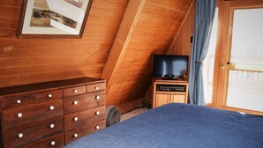 Un dormitorio con un aparador de madera, una cama con manta azul, un televisor sobre un mueble bajo y una ventana con cortinas.