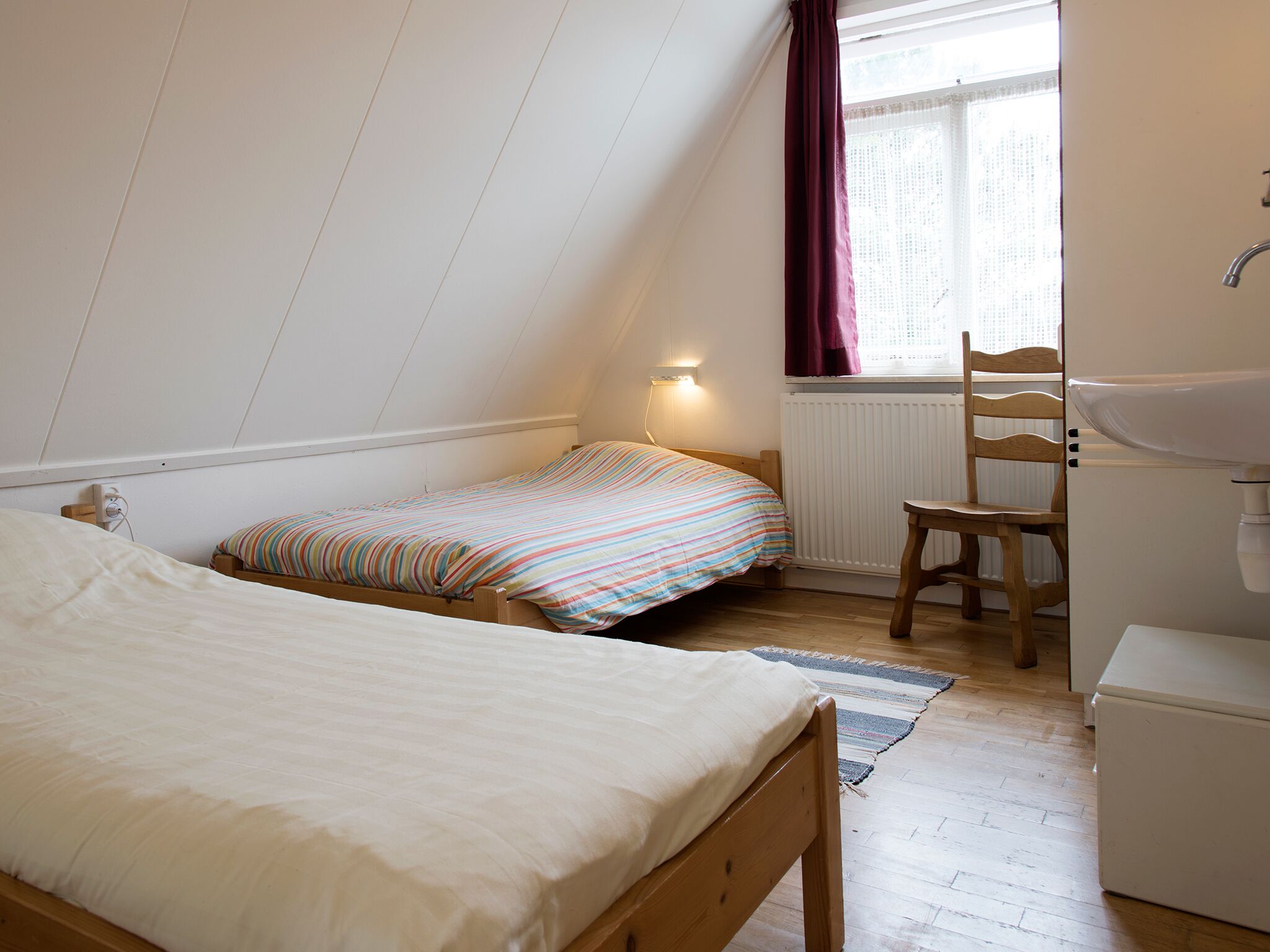 A small, well-lit room with two beds, a chair, a radiator, and a window with curtains.