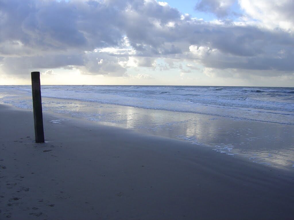 A beach with a wooden post, calm waves, and a cloudy sky.