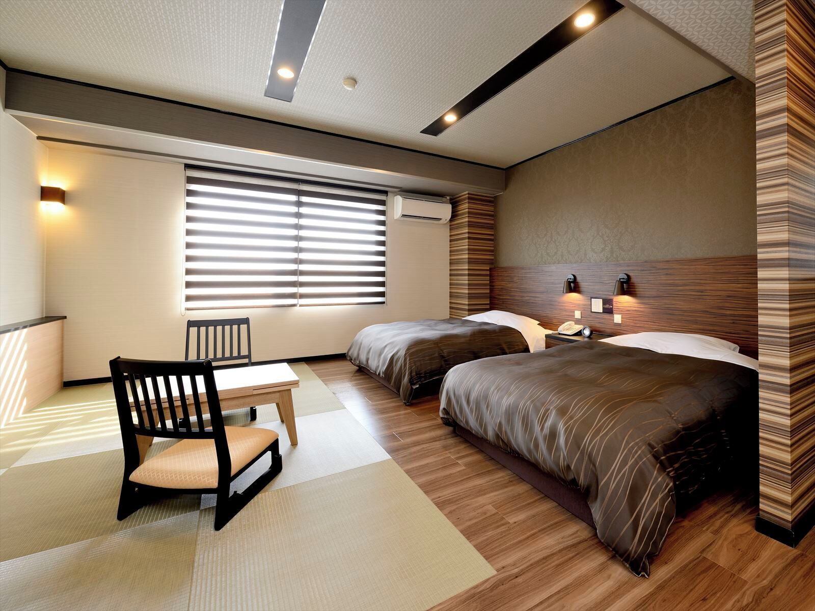 A hotel room with two beds, a desk, a chair, and a large window with blinds.