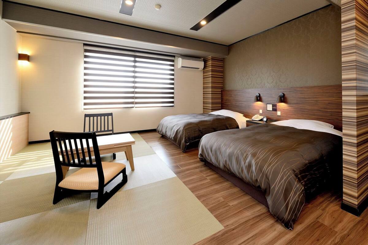 Een hotelkamer met twee bedden, een bureau, een stoel en een groot raam met luxaflex.