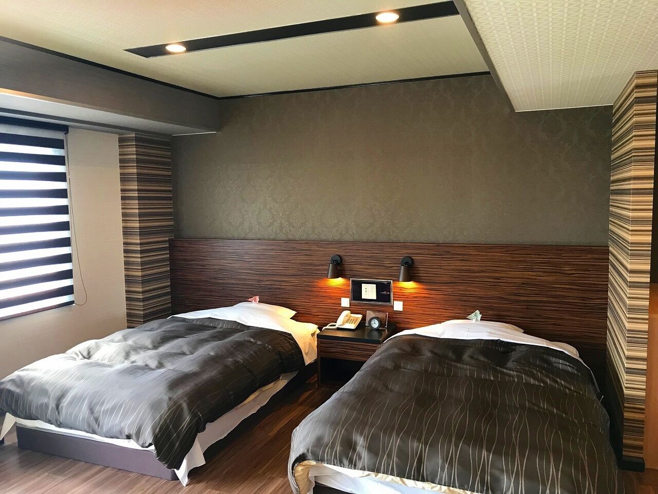 A hotel room with two beds, a bedside table with a phone and a clock, and a wall-mounted light fixture.