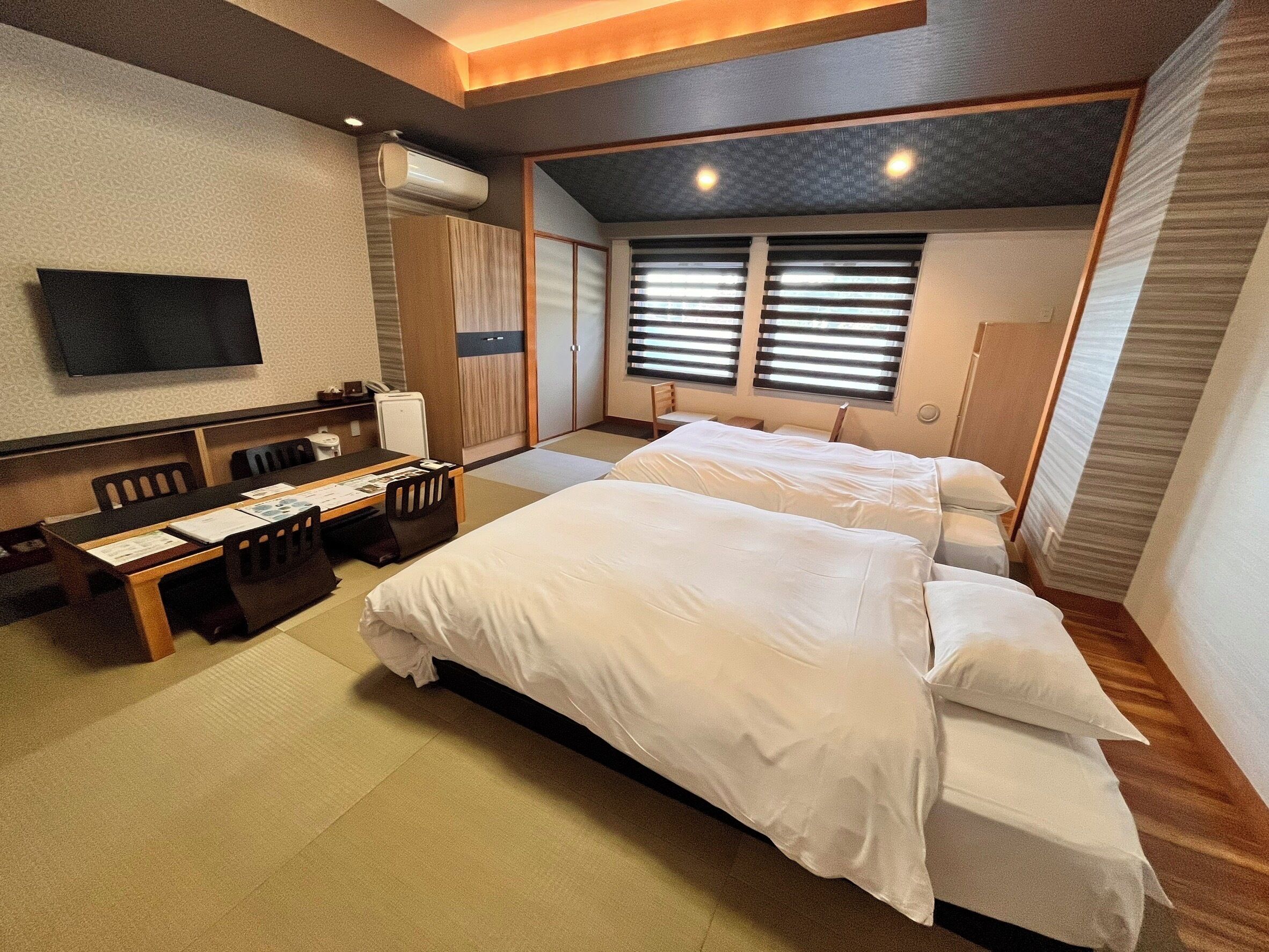 A hotel room with a bed, a desk with chairs, a television, and a window with blinds.