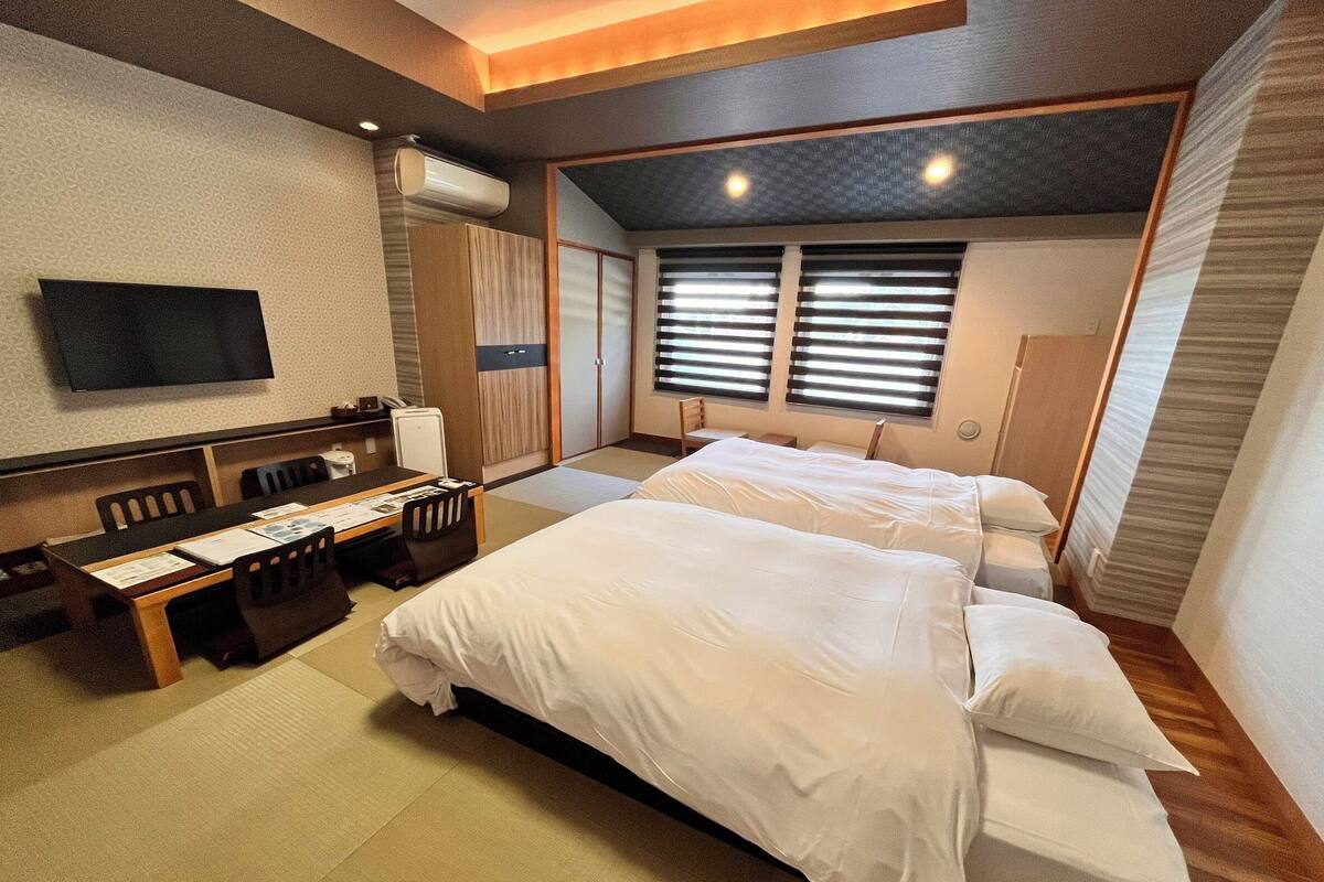 Een hotelkamer met een bed, een bureau met stoelen, een televisie en een raam met jaloezieën.